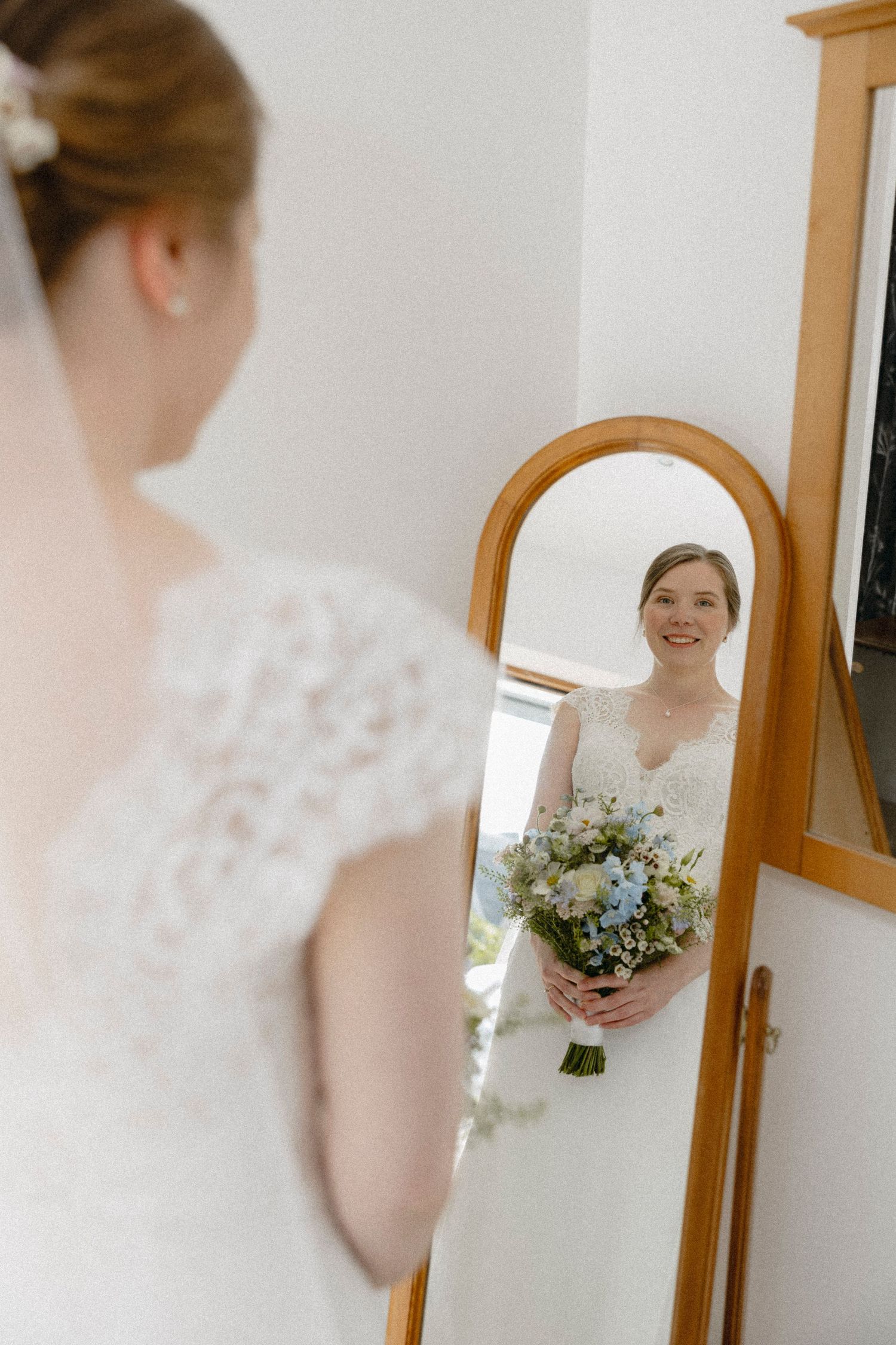 Bride in lace wedding dress holding floral bouquet smiles at her reflection in a wooden framed floor mirror.
