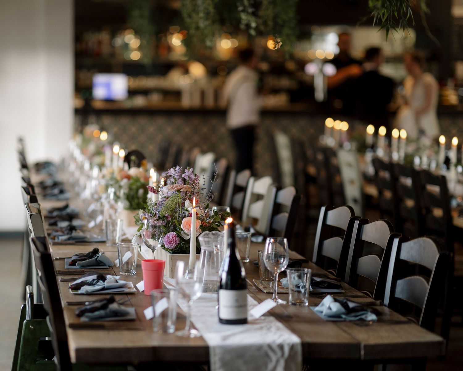 Elegant wedding reception long table with floral centerpieces, candles, wine glasses, and dark wood chairs in a modern venue.