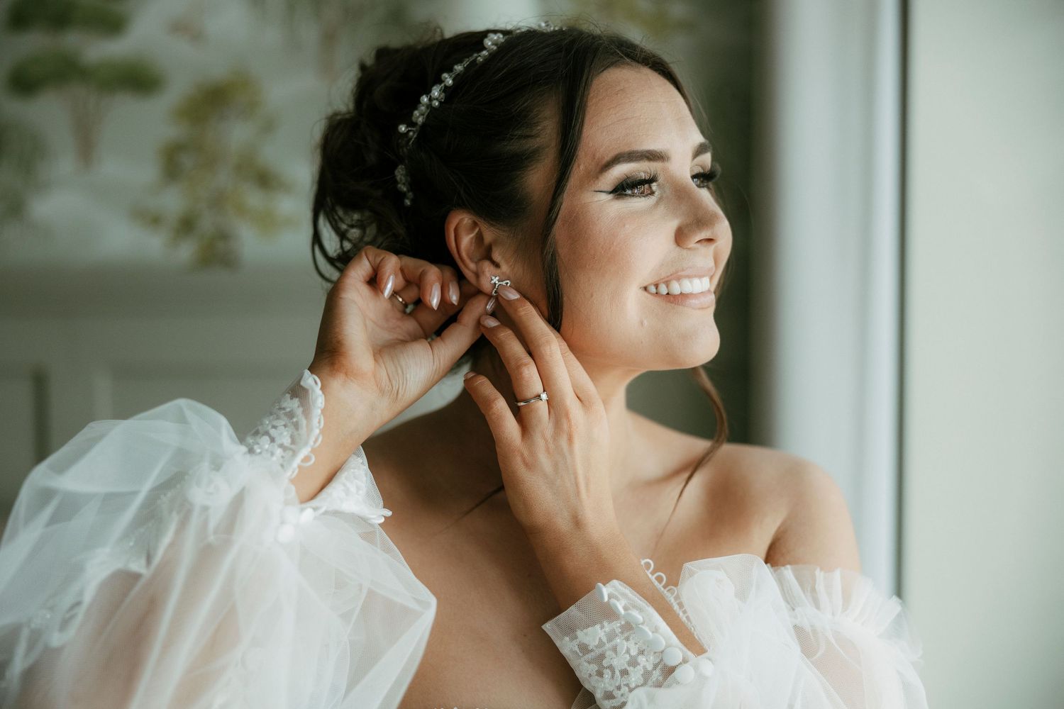 Bride smiling while putting on elegant earrings in wedding dress with sheer sleeves and pearl headband.