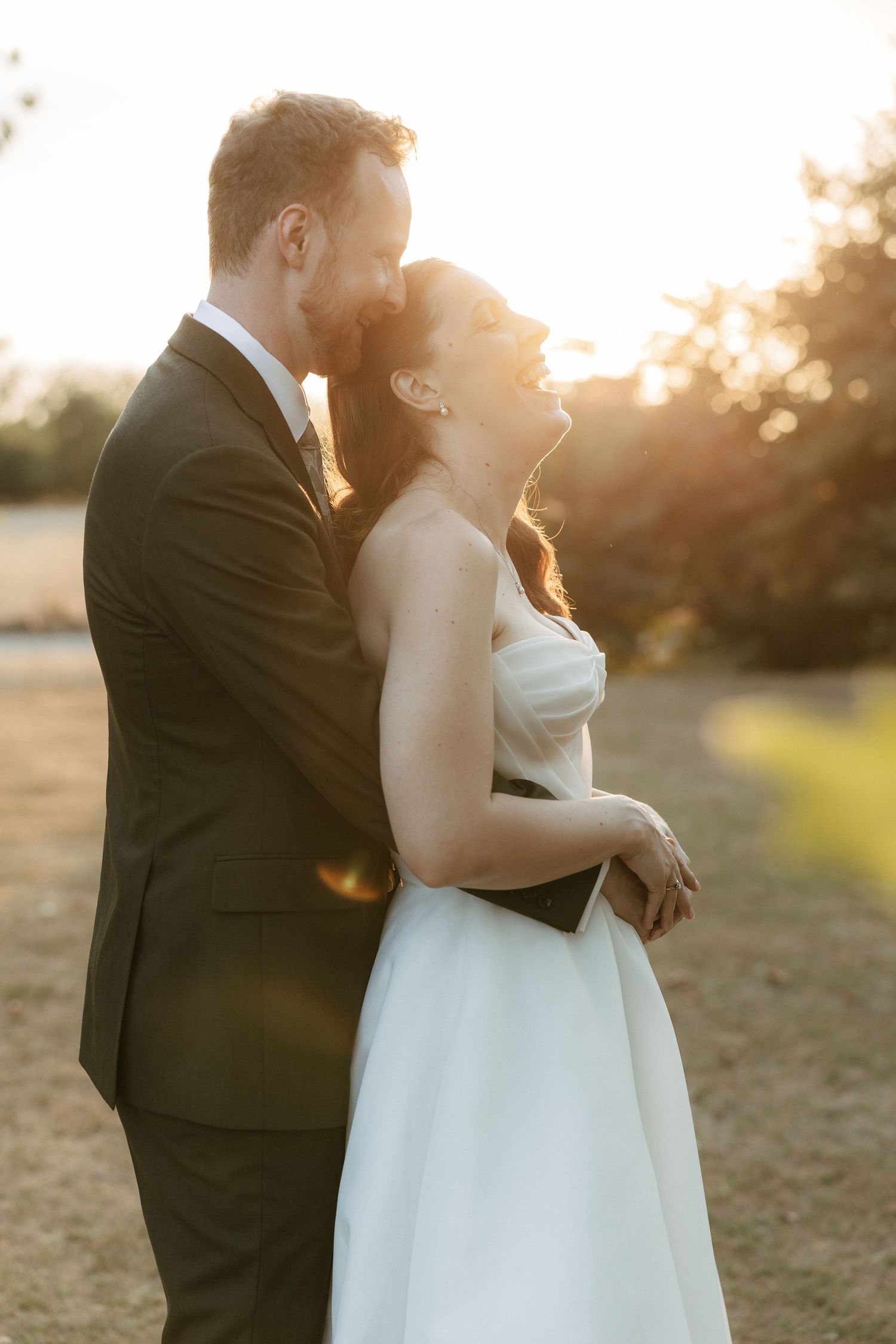 Bride and groom embrace at golden hour sunset, groom kissing bride's cheek as she smiles in white strapless gown.