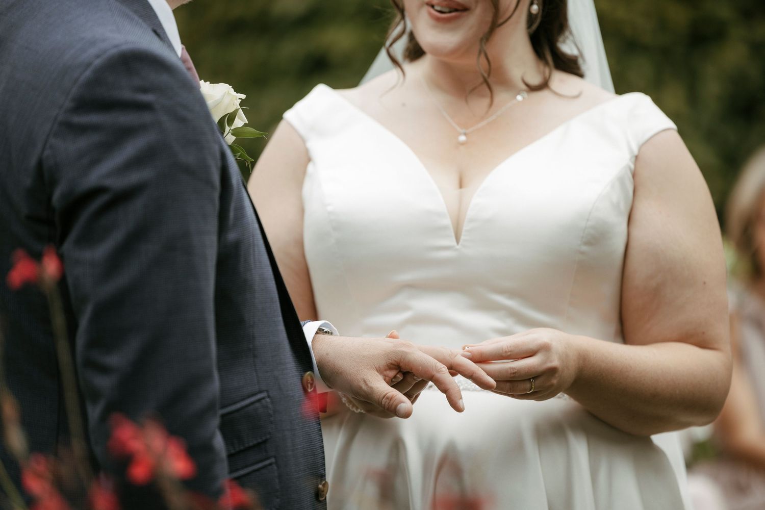 Bride in white off-shoulder gown placing wedding ring on grooms finger during outdoor ceremony.