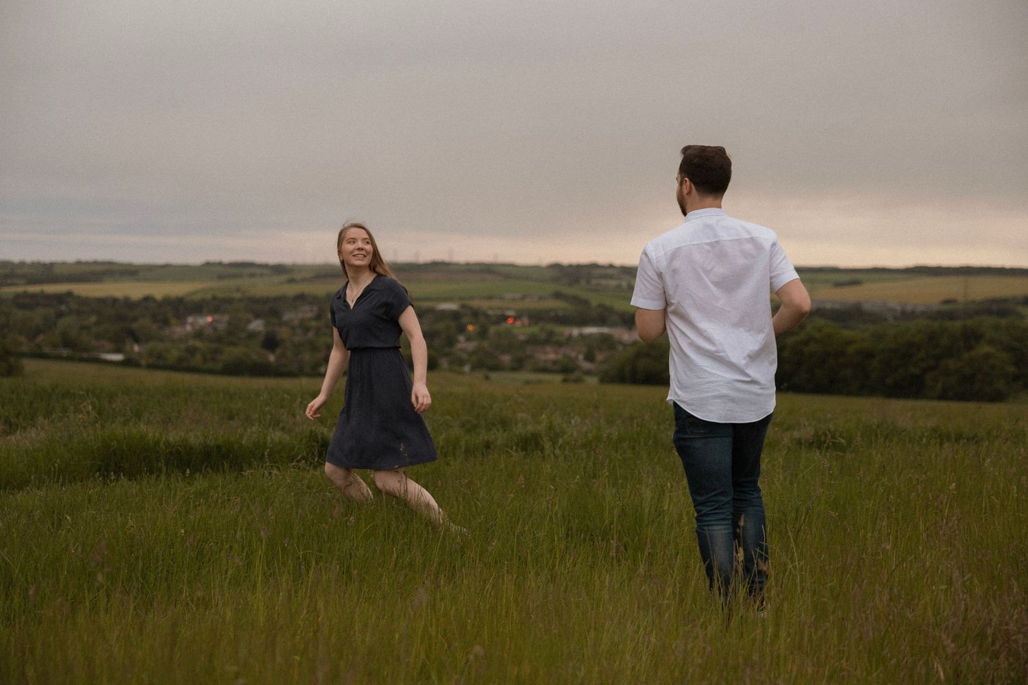Two people playfully running through a grassy meadow with rolling hills in the background.