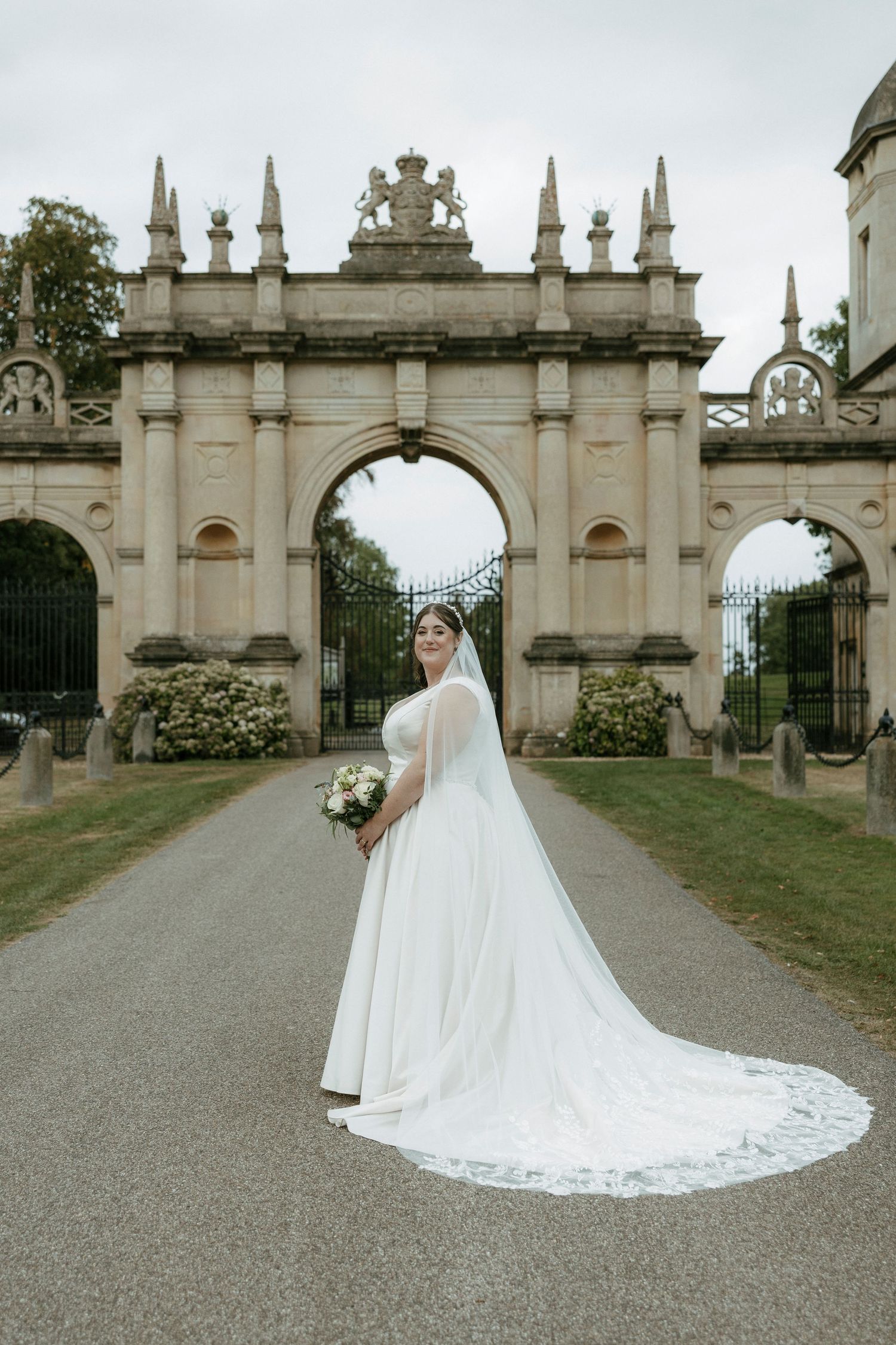 Bride in elegant white gown and long veil holding bouquet before ornate stone arch gateway.