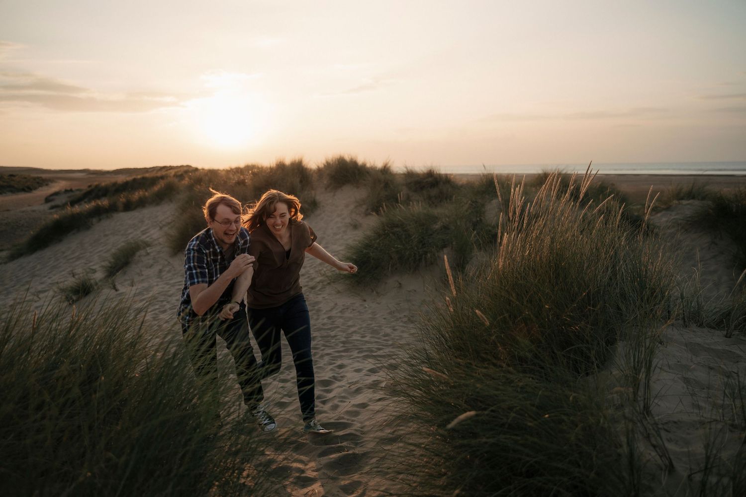 Couple laughing and running through sandy beach dunes at sunset.