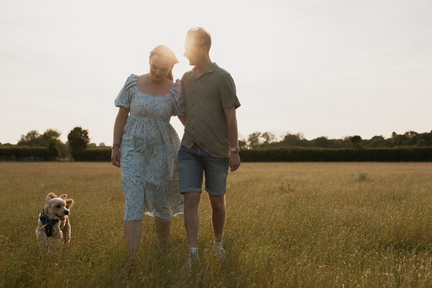 Couple walking with a dog in a golden sunset field, woman in floral dress and man in casual shorts.