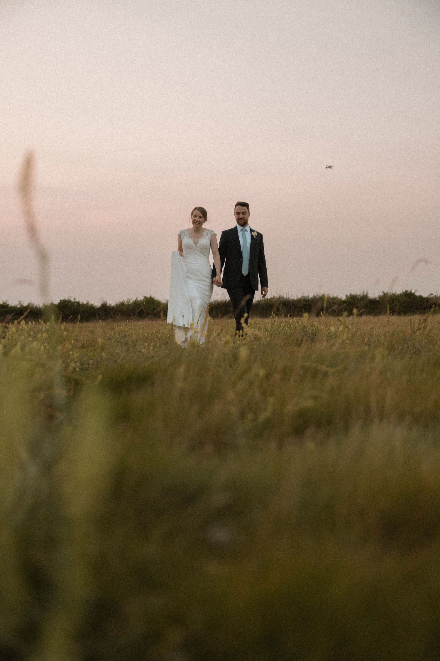 A bride and groom walk hand in hand through a lush green field at sunset on their wedding day.