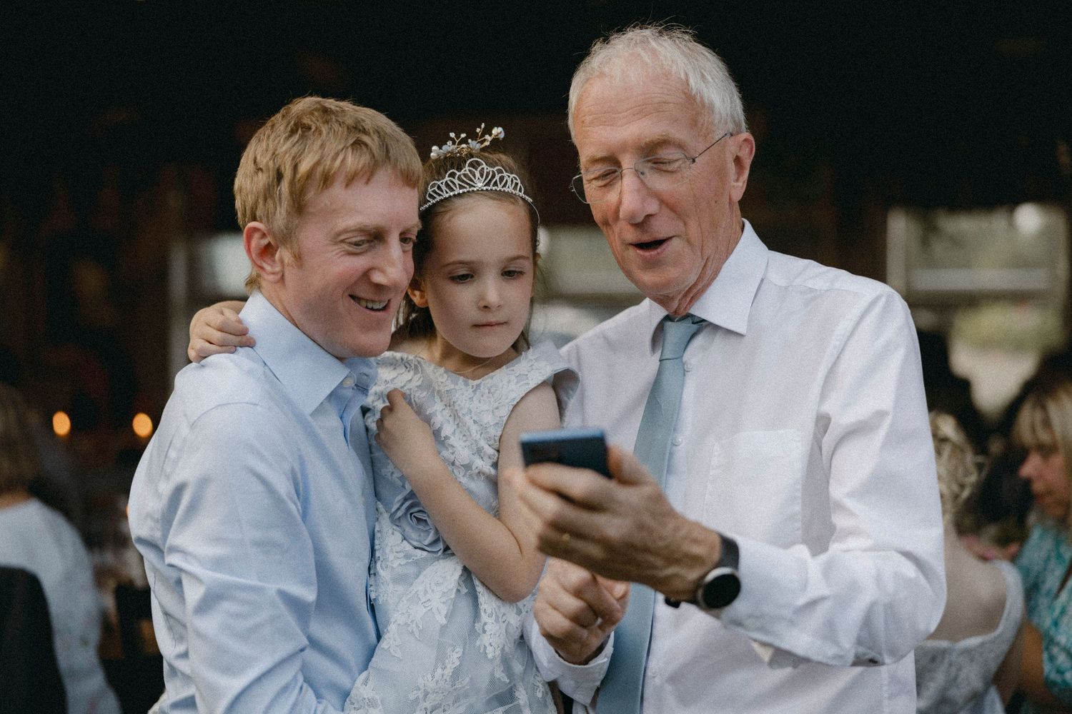 Two men and a young girl in a tiara smile while viewing a smartphone at a wedding celebration.