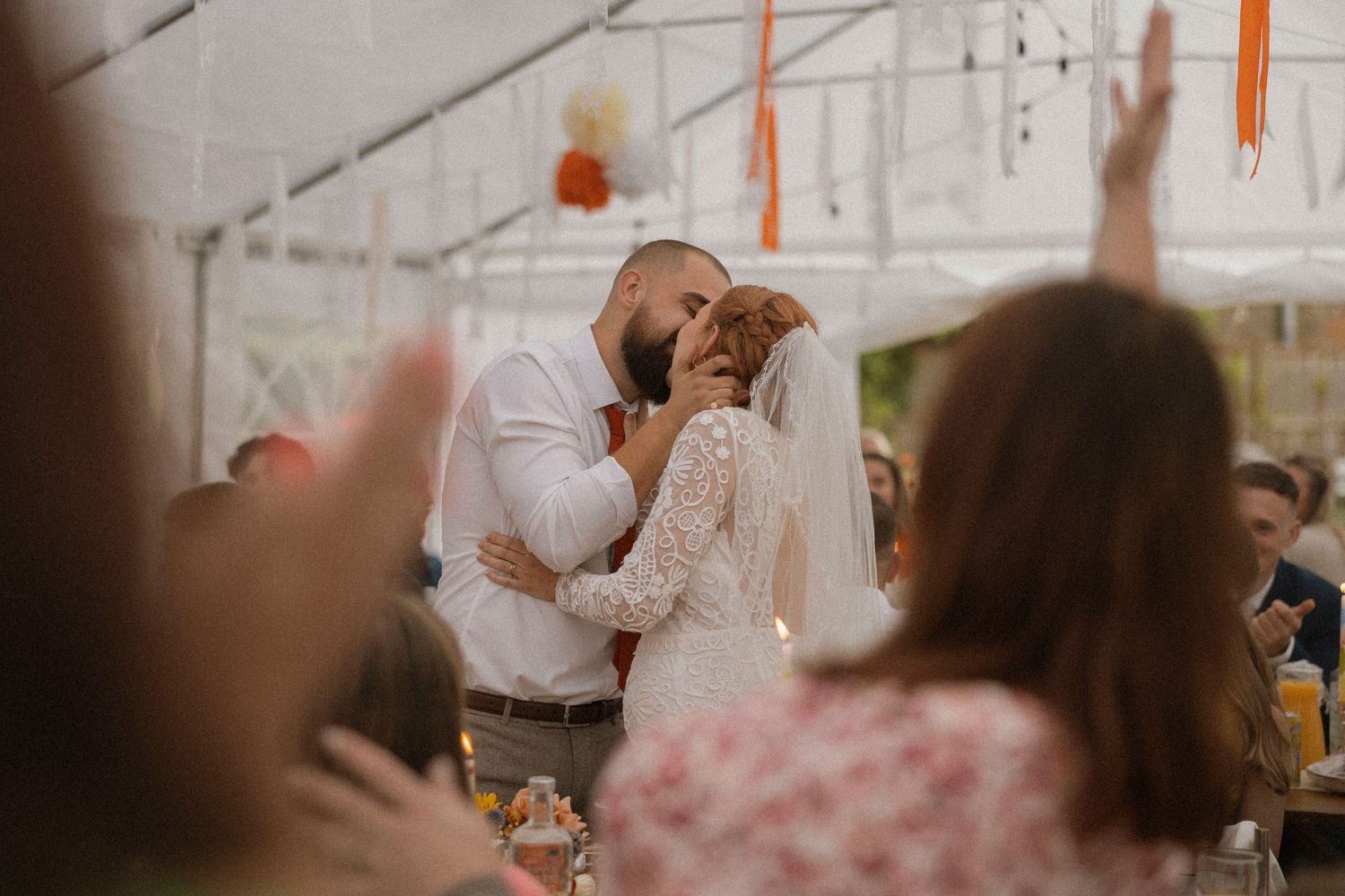 Bride and groom share a romantic kiss at outdoor wedding reception under white tent with guests celebrating.