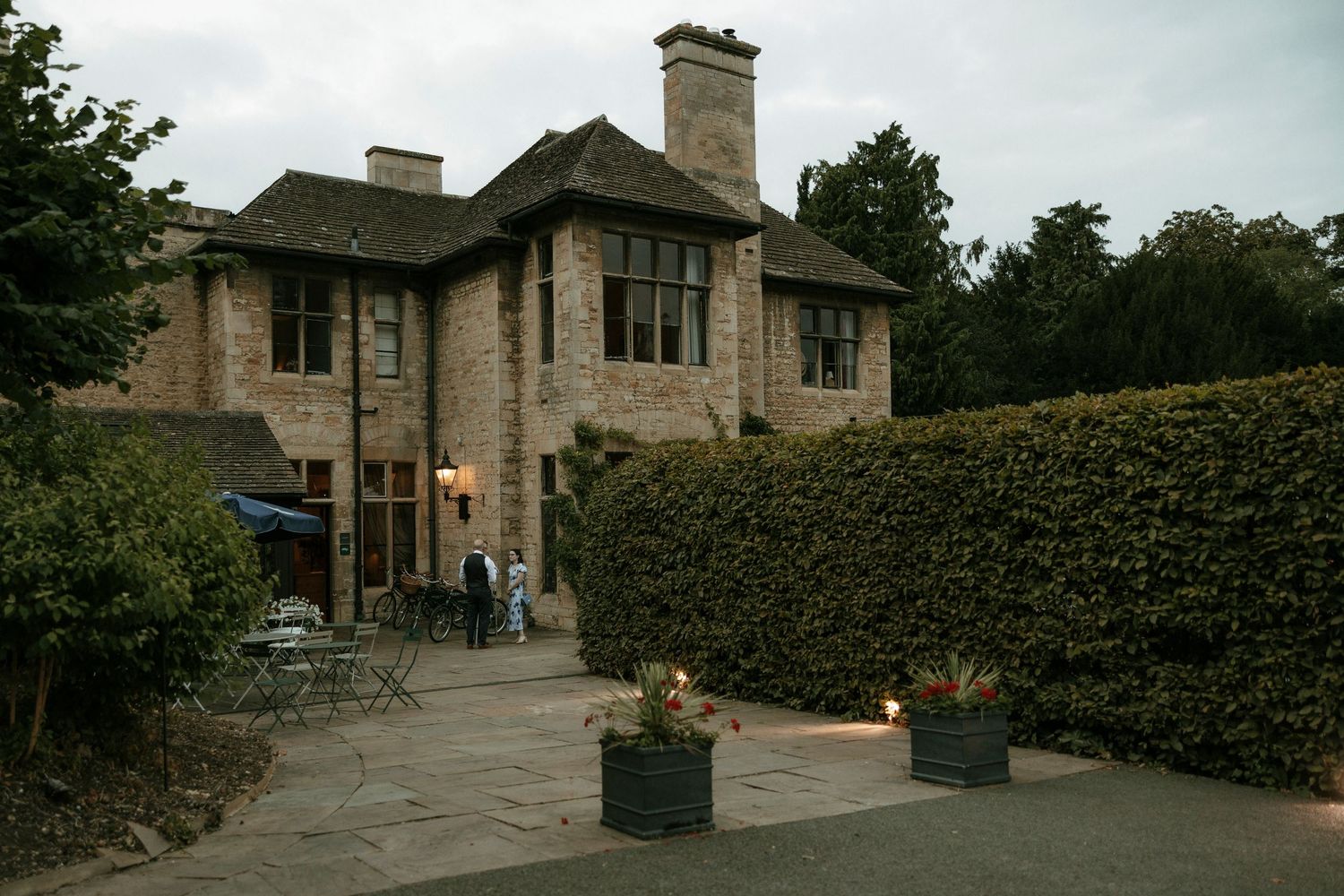 Stone manor house with patio, potted plants, and hedgerow at dusk, with guests gathered outside.