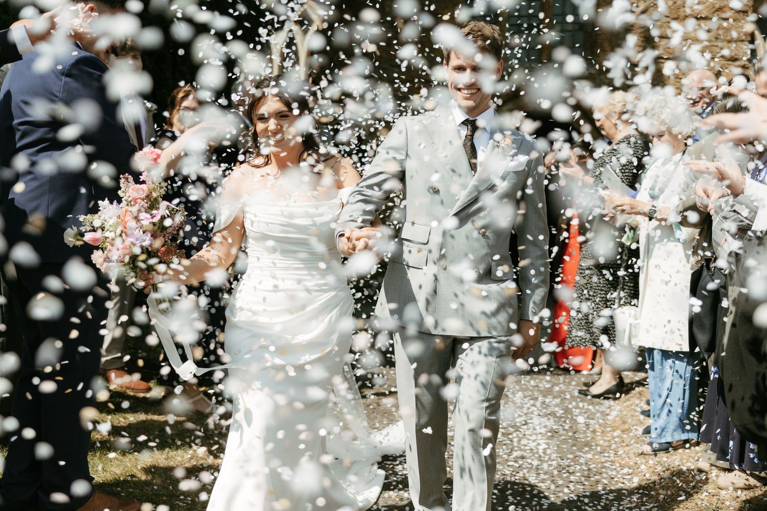 Bride and groom walking through confetti shower at outdoor wedding ceremony celebration.