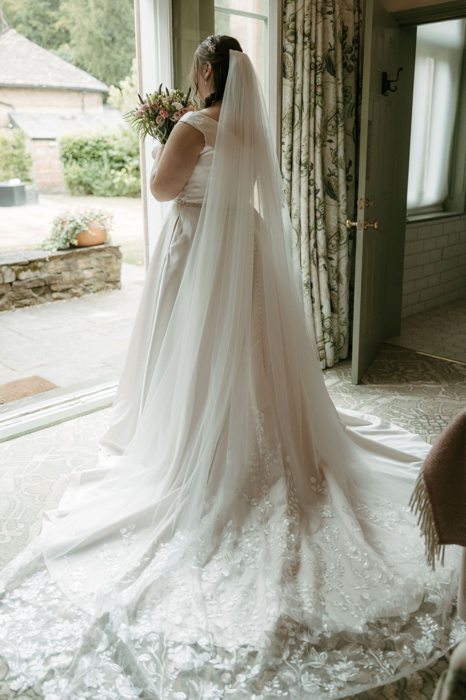 Bride in elegant lace wedding gown with long veil holding bouquet by window.