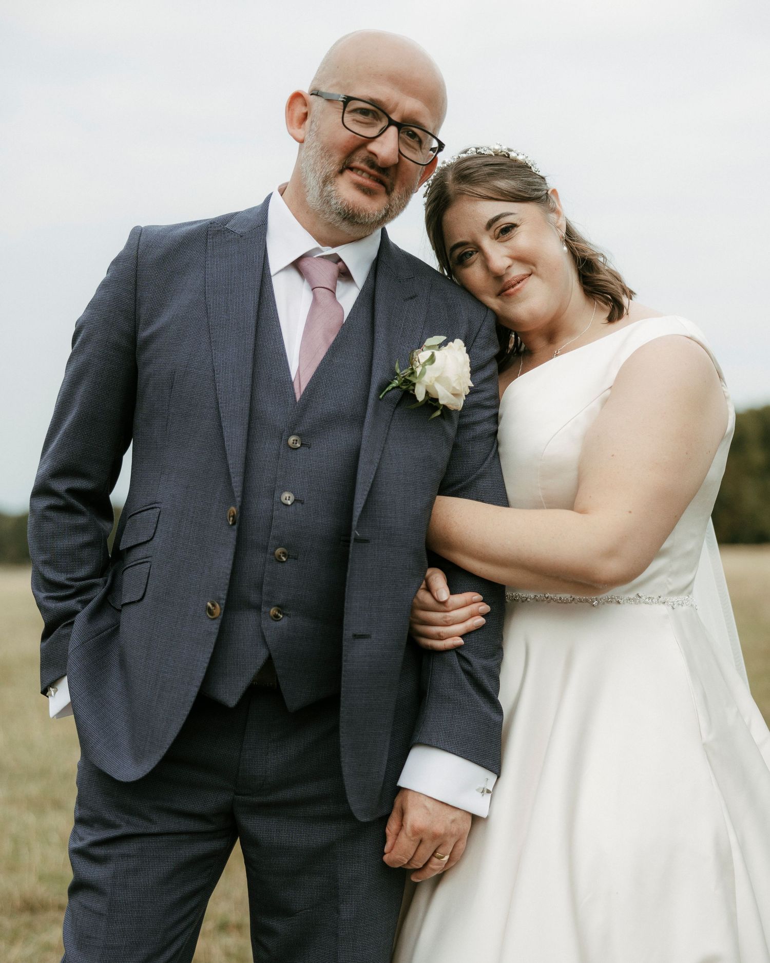 Smiling bride and groom posing together in field, groom in grey suit, bride in white gown.