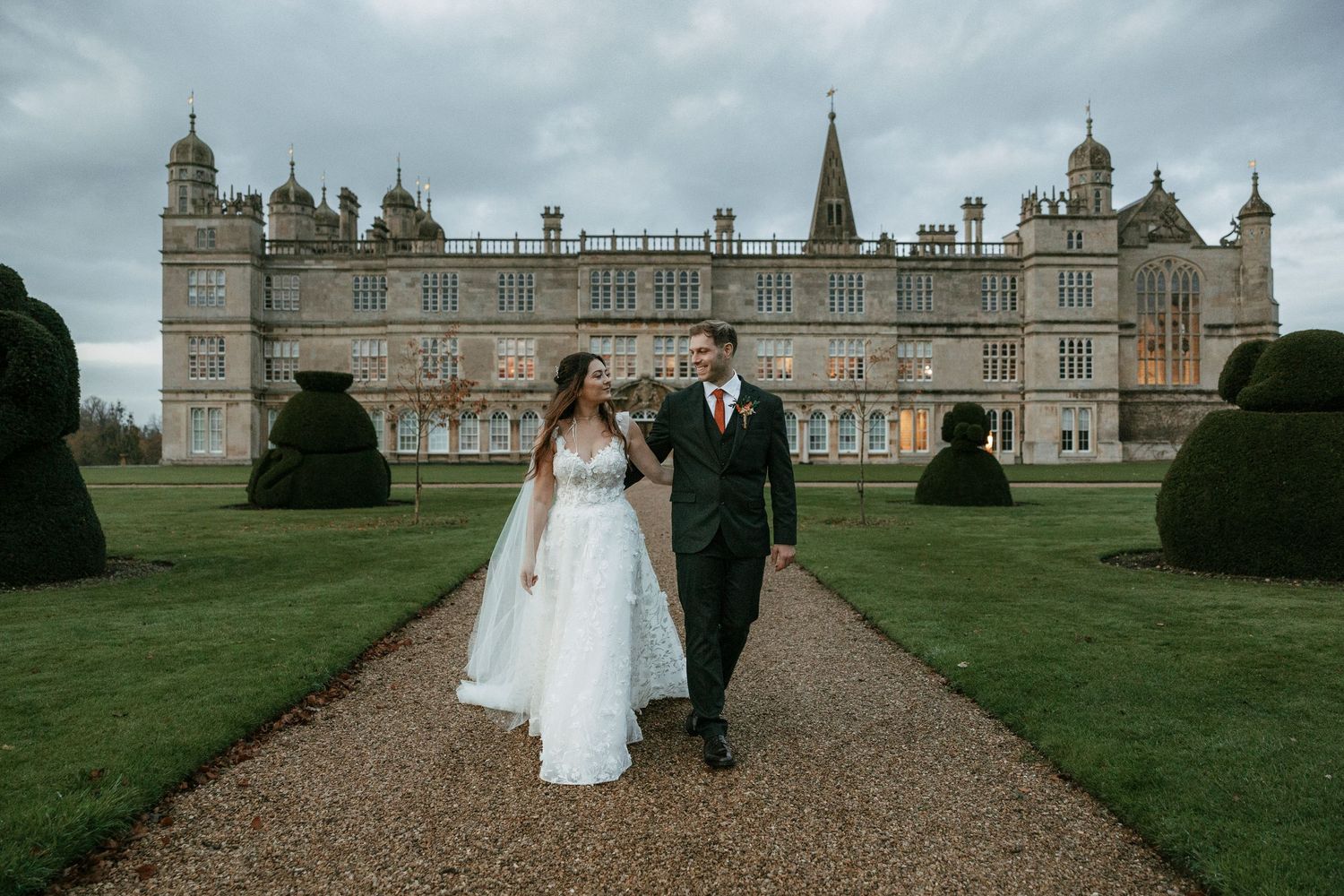 Bride and groom walking hand in hand on garden path before grand English castle wedding venue.