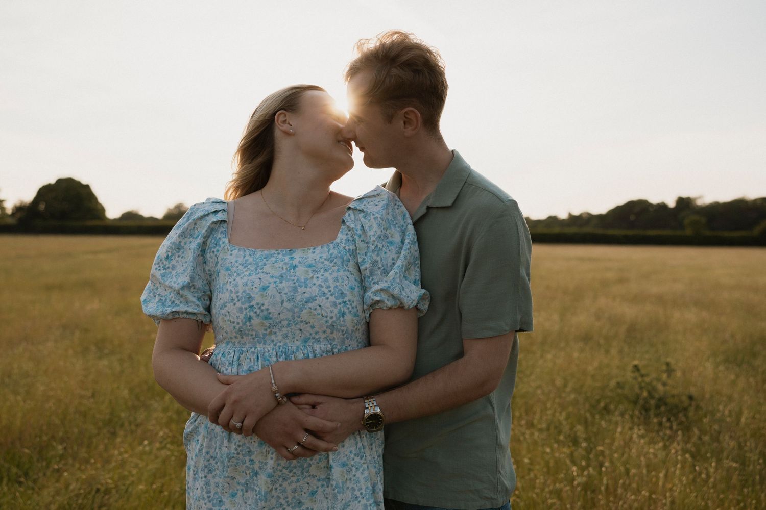 Couple sharing a tender moment in a sunlit field, woman in floral dress with baby bump, golden hour backdrop.