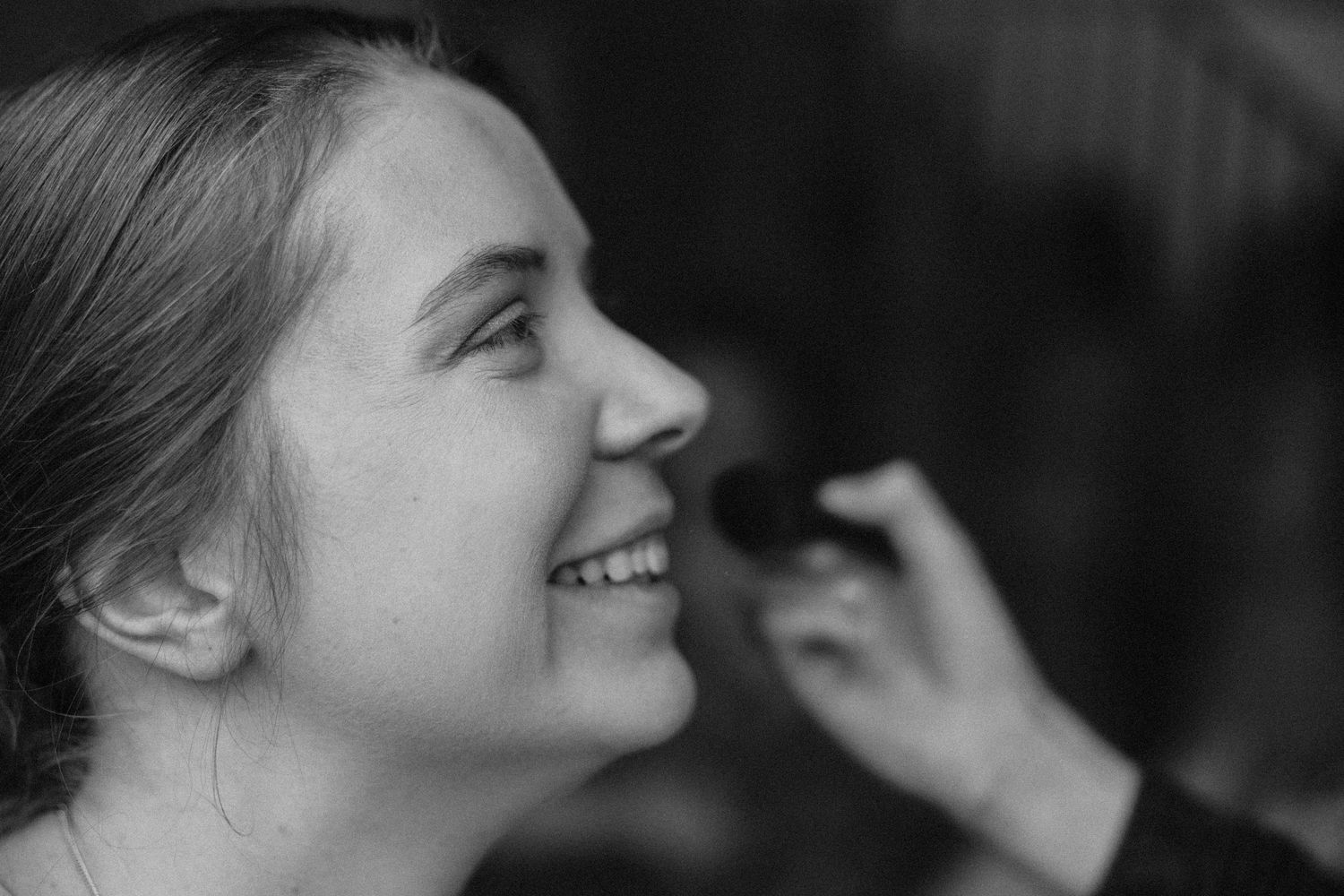 Young woman smiling as makeup artist applies cosmetics, captured in black and white photography.