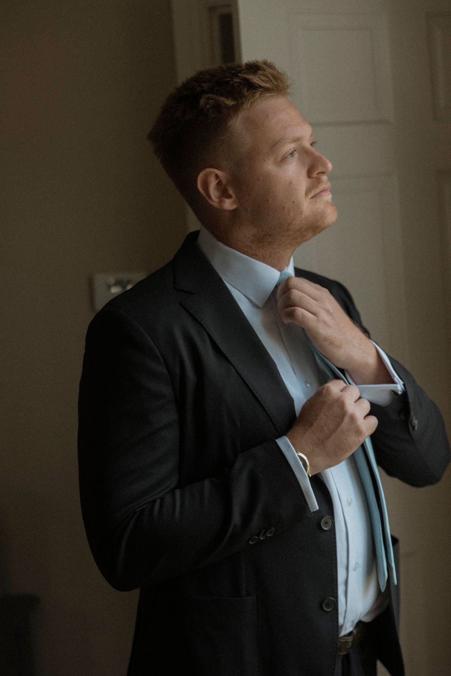 A groom in a dark suit adjusts his light blue tie while gazing upward in soft natural light.