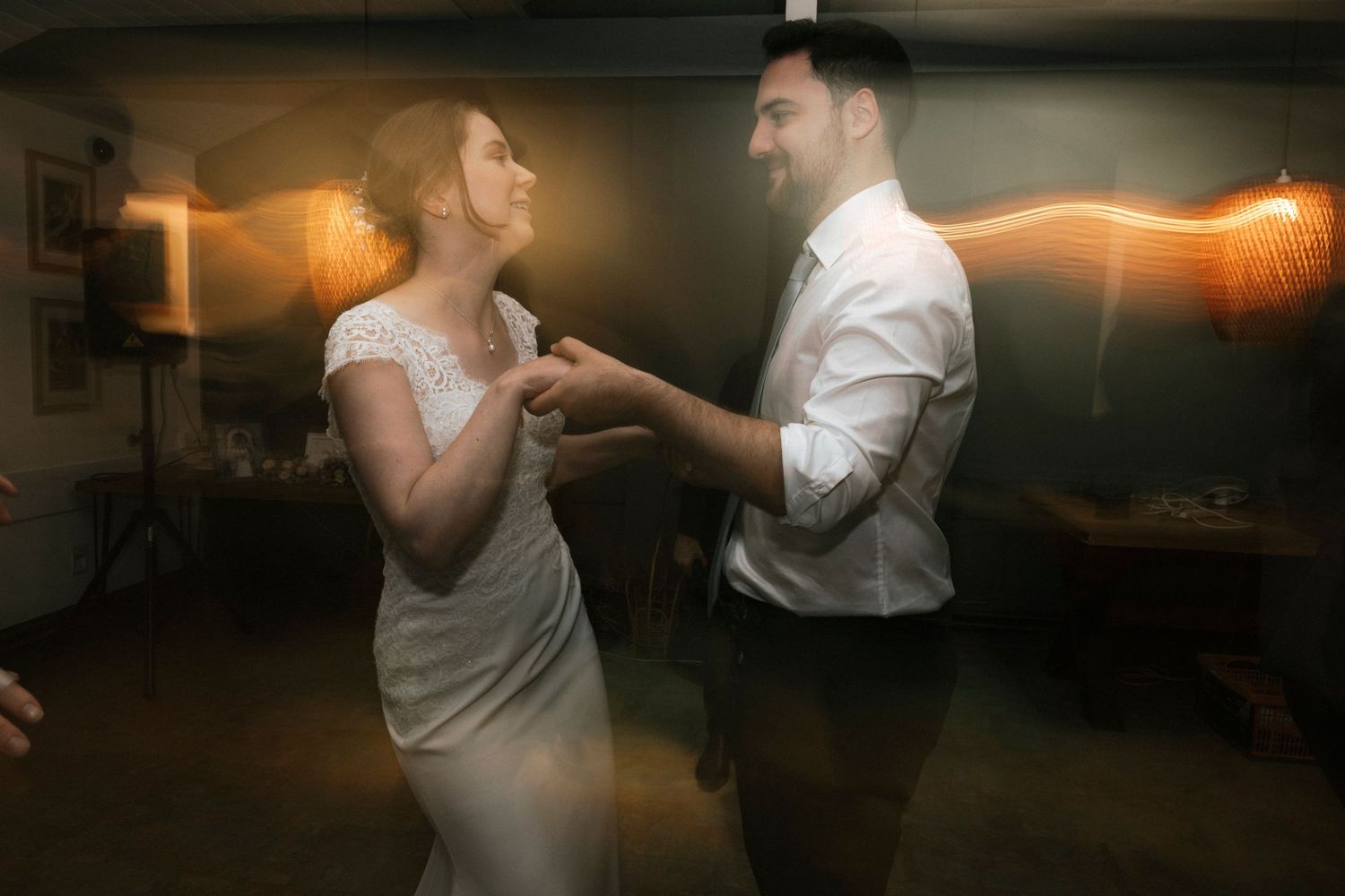 A bride and groom share their first dance, holding hands and smiling in warm, romantic lighting.