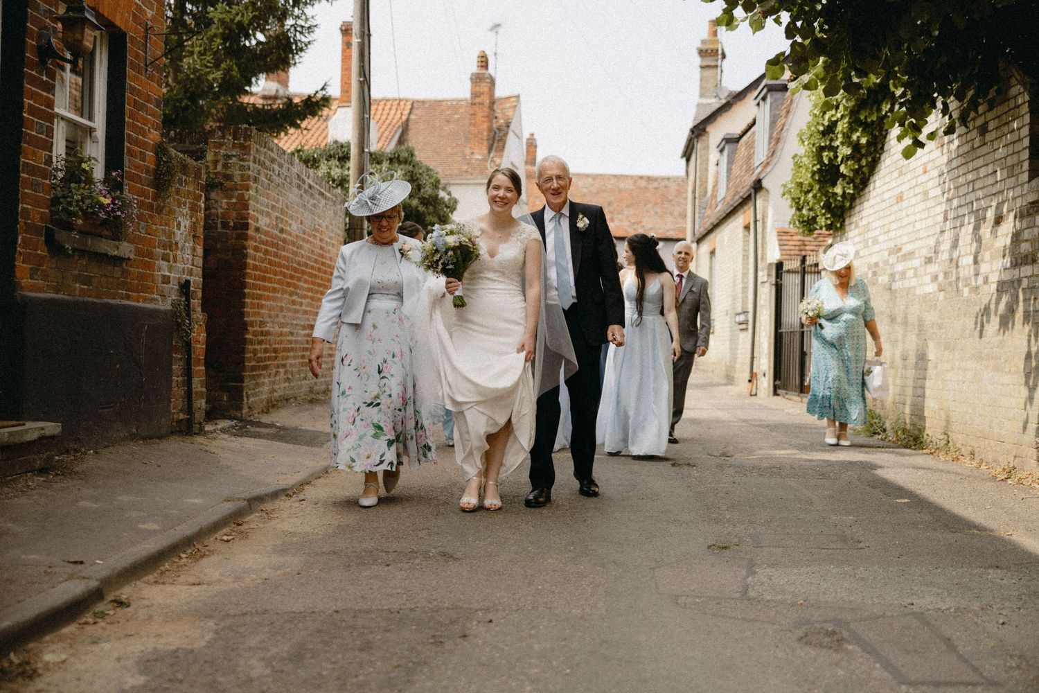 Bride in white gown walks with family through a charming English village street on her wedding day.