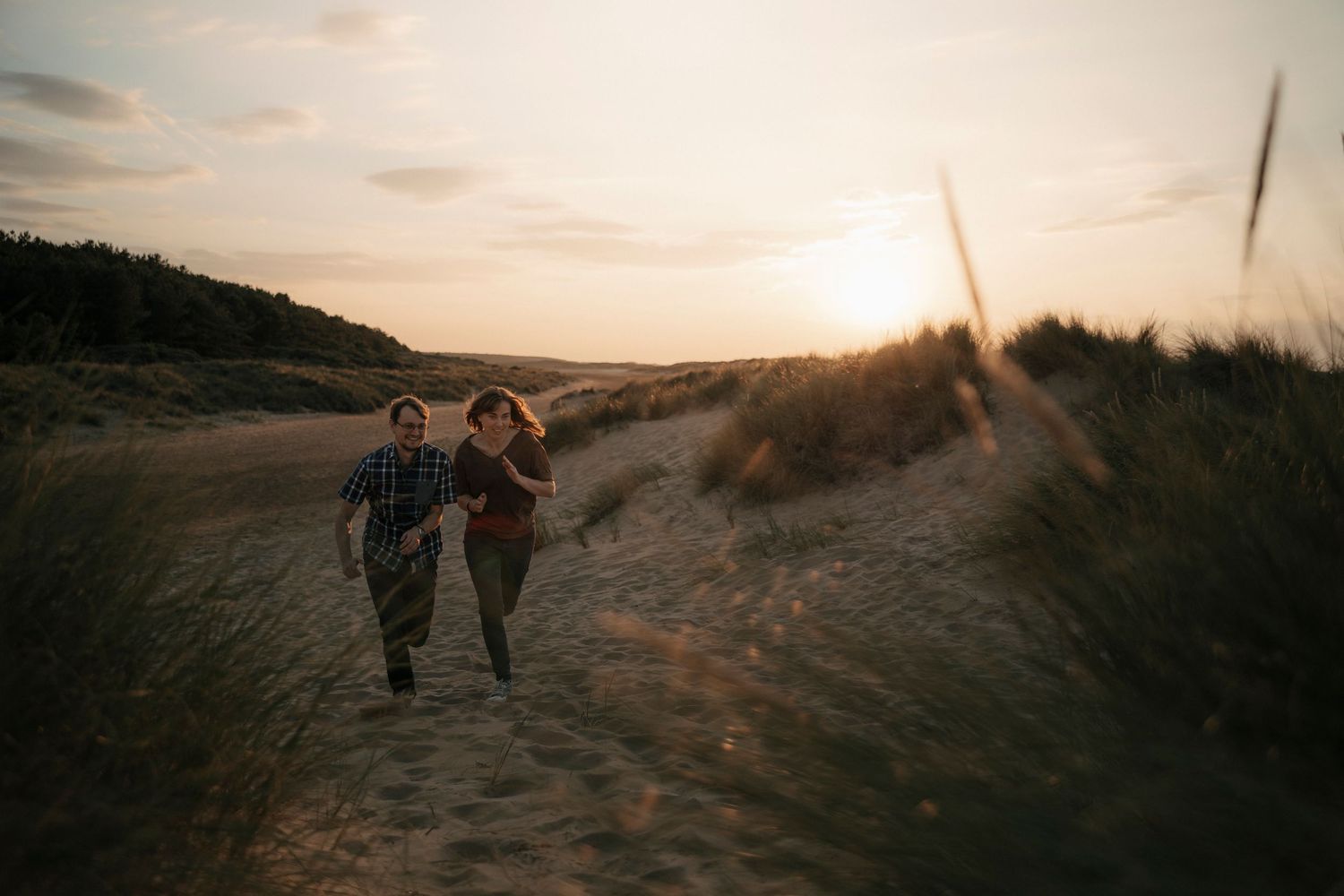 Couple running hand-in-hand through sandy dunes at golden sunset, surrounded by tall grass on a scenic beach.