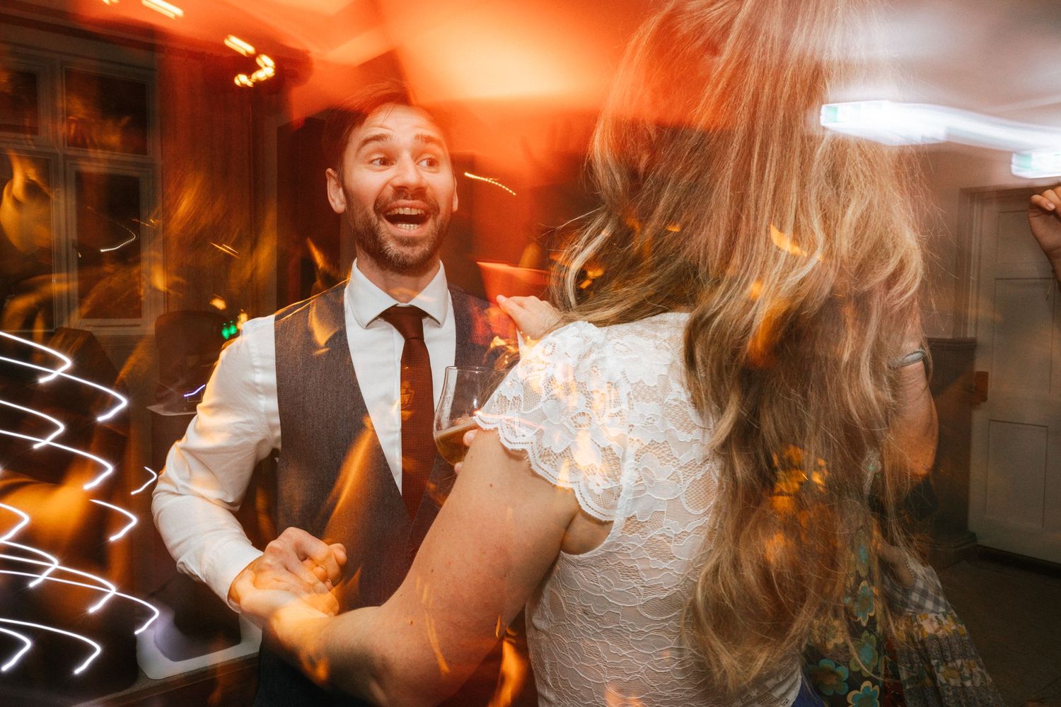 Joyful groom dancing with bride at wedding reception, warm bokeh lights glowing in background.