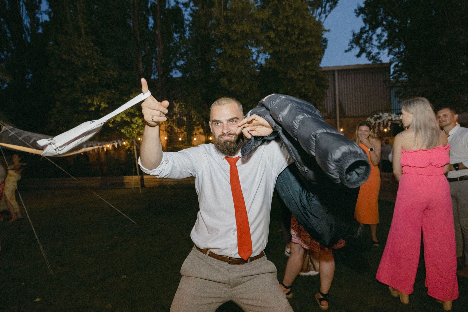 Bearded man in white shirt and red tie celebrating outdoors at evening party, giving thumbs up.