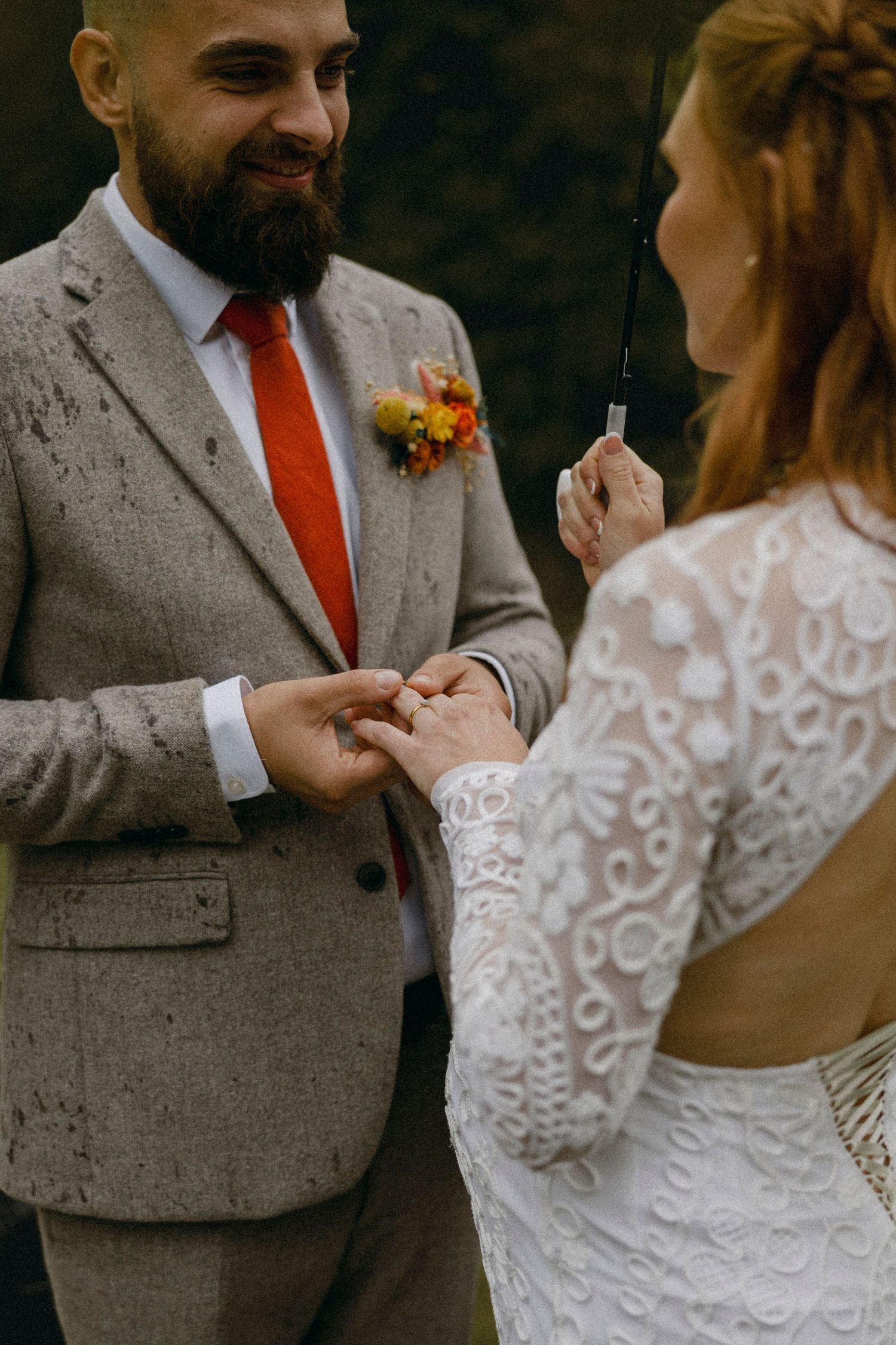 Bride in lace gown places wedding ring on groom's finger during outdoor ceremony, groom wearing gray suit and red tie.