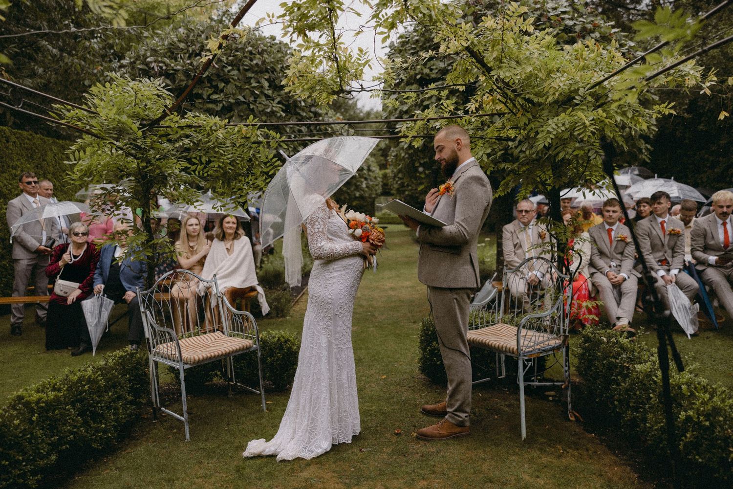 Outdoor garden wedding ceremony with bride in lace gown holding umbrella and groom in tan suit before seated guests.