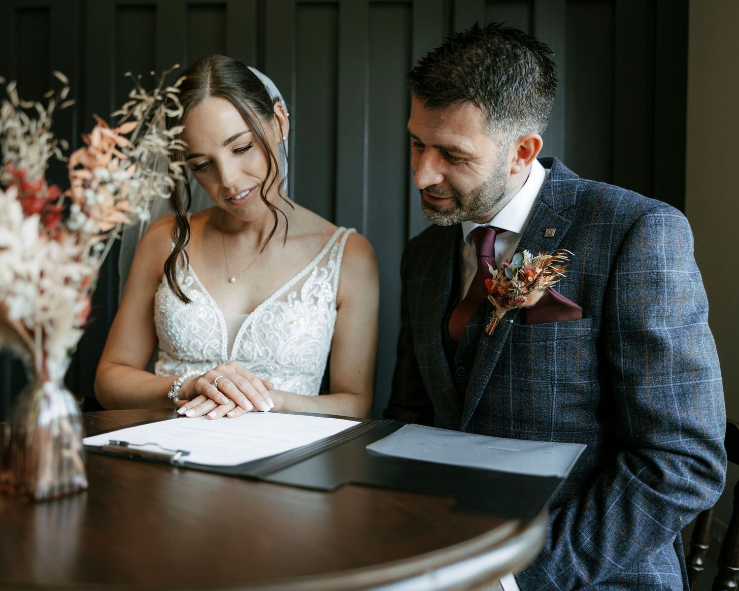 Bride and groom signing wedding register at wooden table, groom in plaid suit with floral boutonniere.