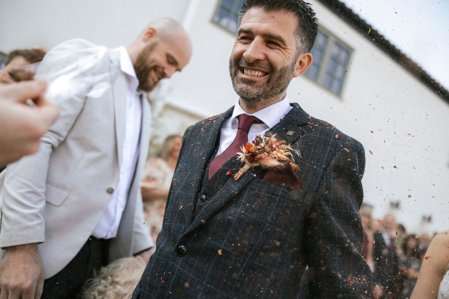 Smiling groom in tweed suit with floral boutonniere celebrates with confetti at outdoor wedding ceremony.