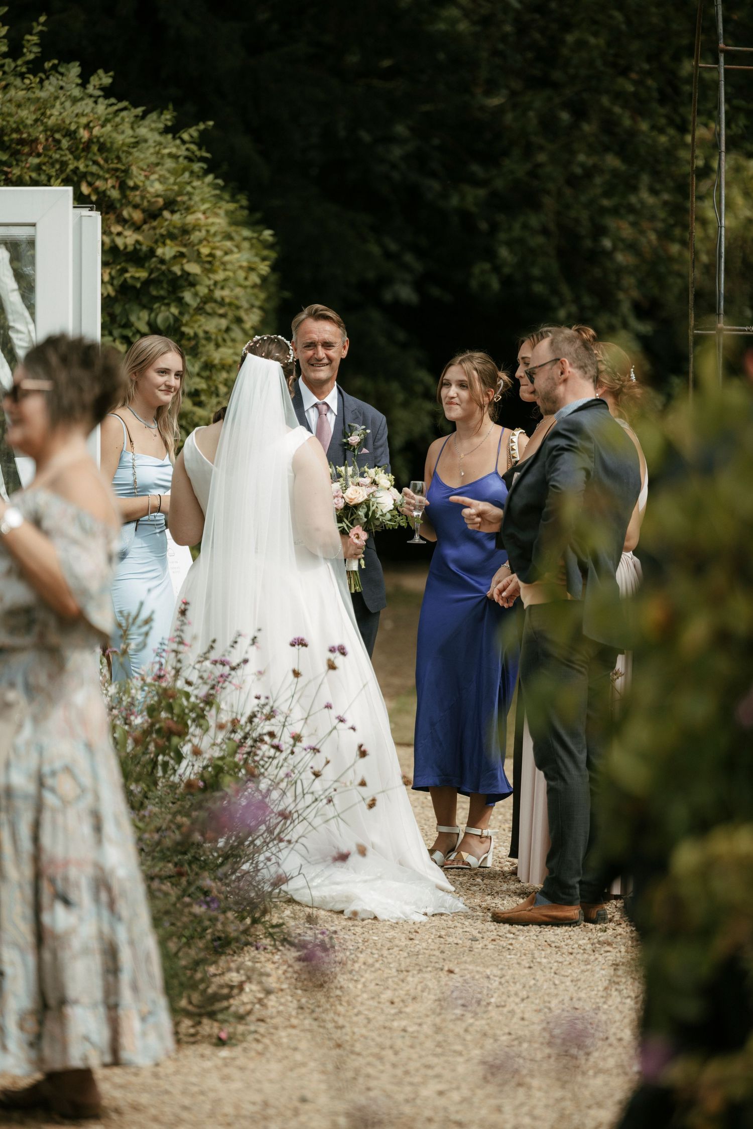Bride in white gown and veil stands with groom and guests outdoors at a garden wedding ceremony.