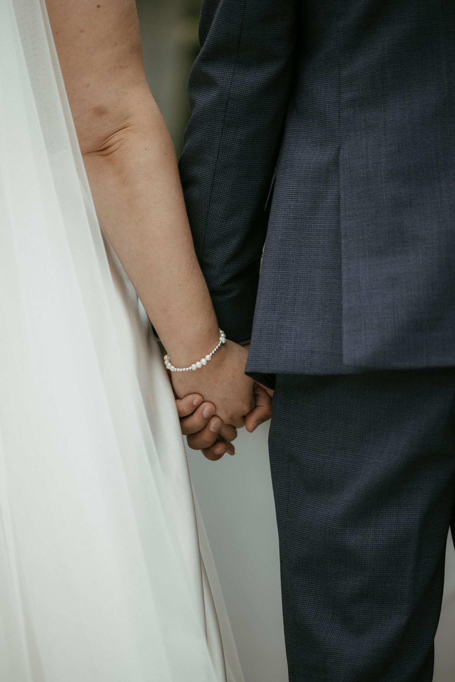 Close-up of bride and groom holding hands, bride wearing pearl bracelet with white gown and groom in navy suit.