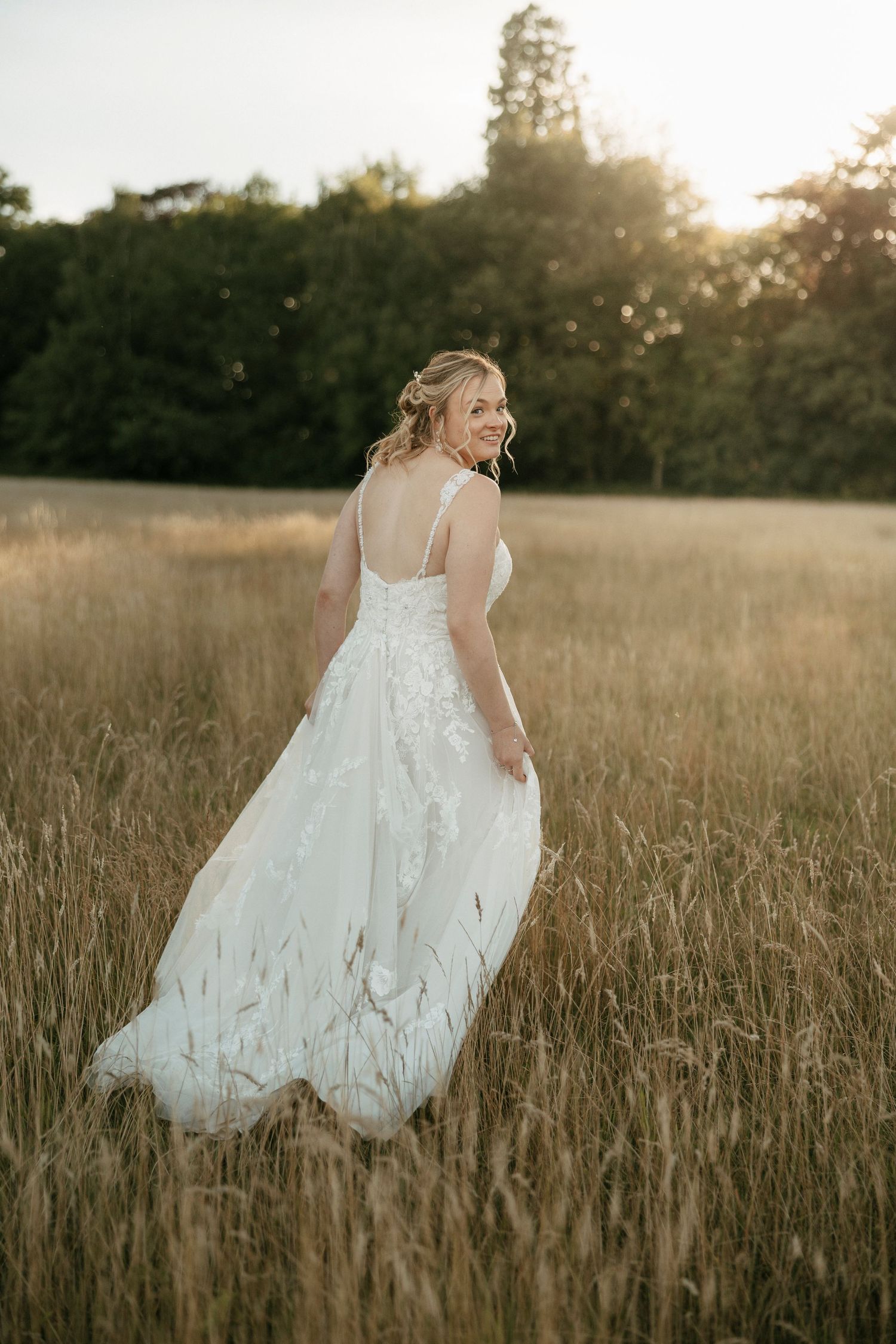Bride in white floral gown twirling in golden field at sunset with trees in background.