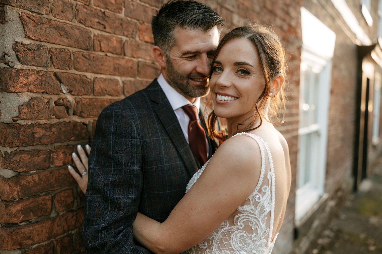 Wedding couple poses intimately against weathered brick wall with natural lighting.