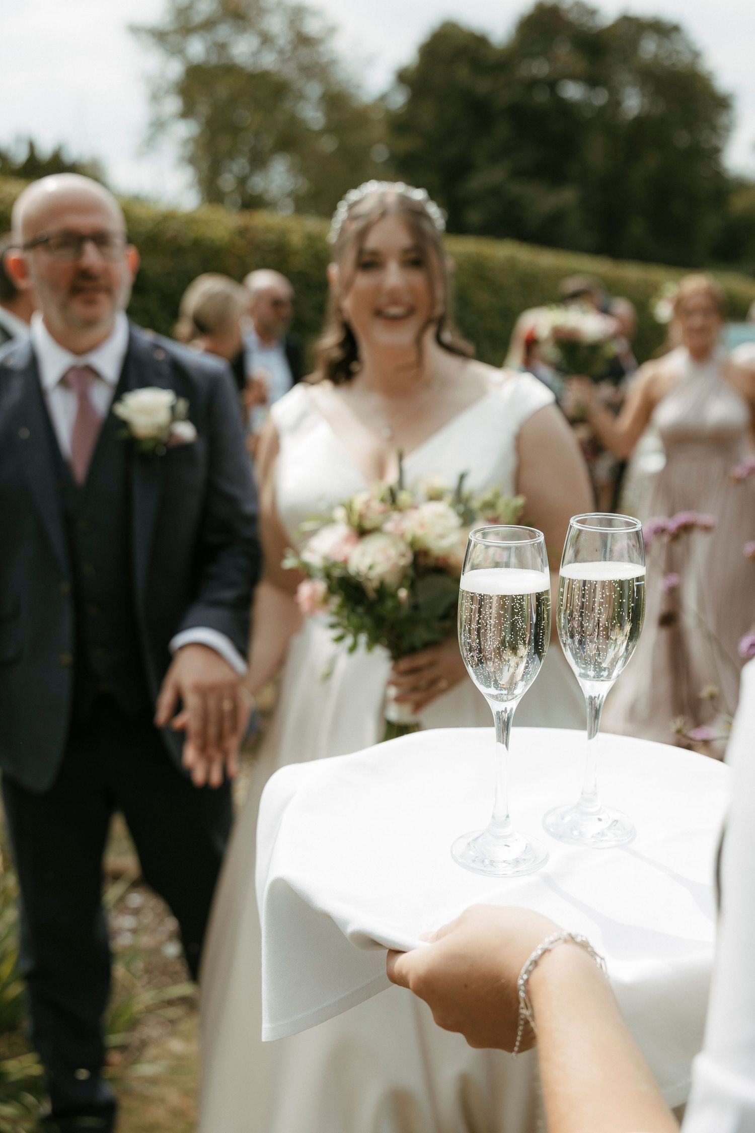 Two champagne flutes on white tray with smiling bride and groom approaching in background at outdoor wedding.