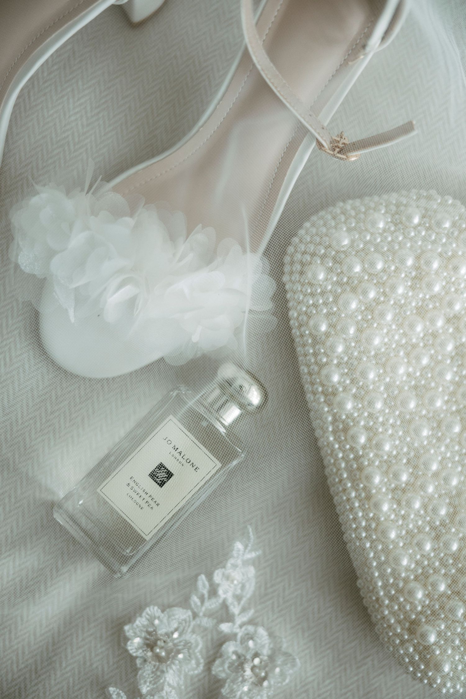 Elegant bridal flat lay with Jo Malone perfume, pearl clutch, white heels, floral garter, and lace accessories.