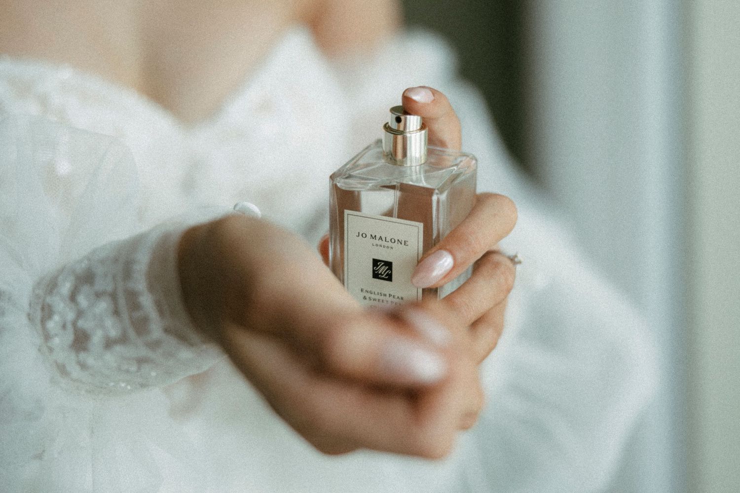 Bride in lace dress holding a Jo Malone perfume bottle on her wedding day.