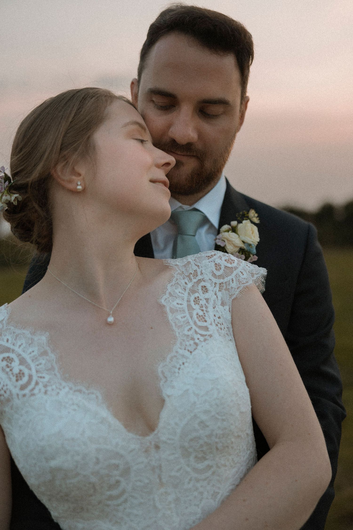 Bride in lace gown and groom in dark suit share a romantic sunset moment, with him gently kissing her cheek outdoors.