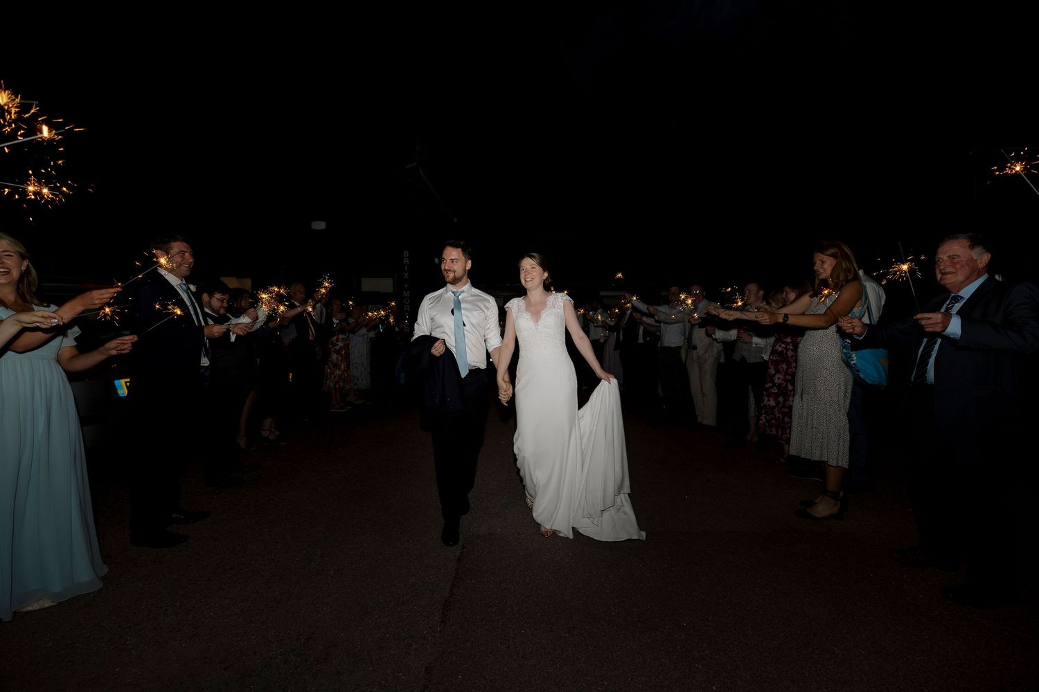 Bride and groom run through sparkler exit tunnel at night during outdoor wedding reception celebration.