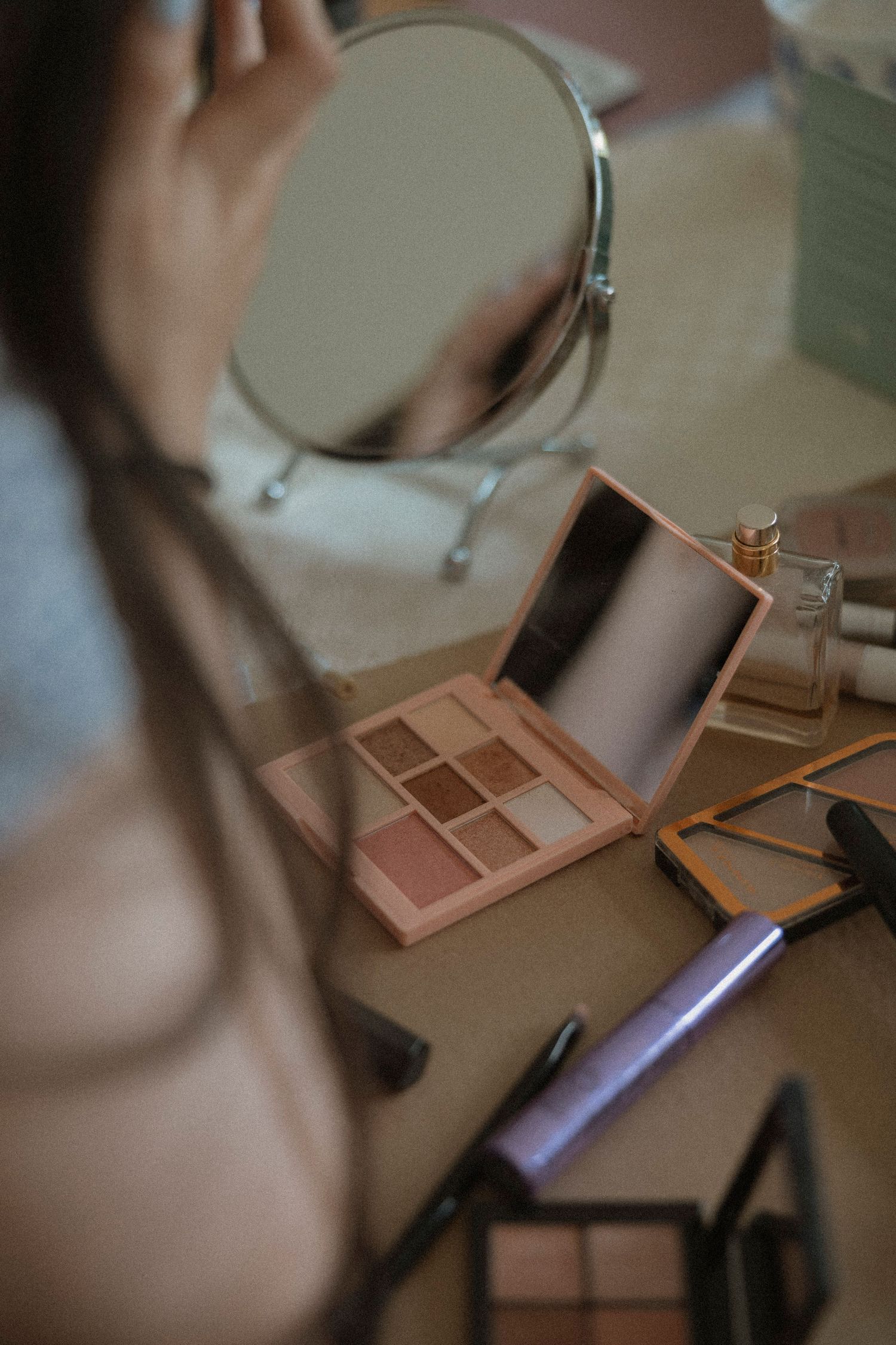 Woman applying makeup at vanity with eyeshadow palette, brushes, and mirror on table.