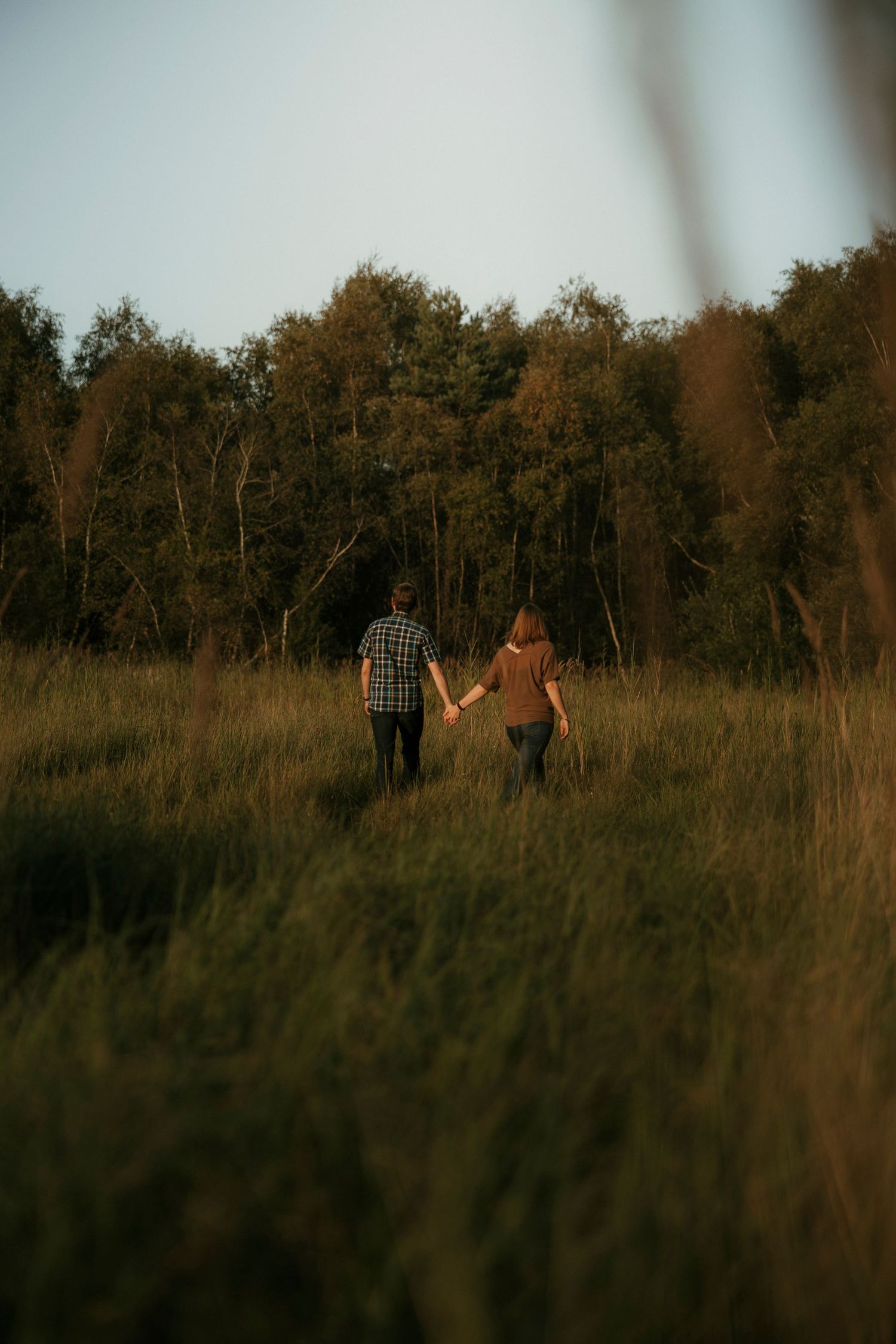 Couple holding hands walking through tall green grass field toward forest trees at golden hour sunset.