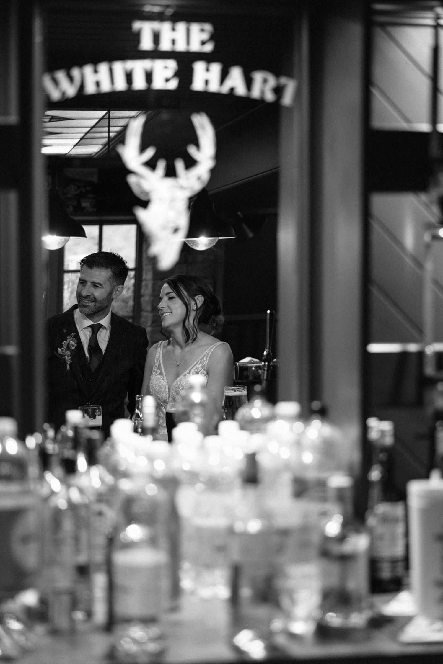 Black and white photo of couple at The White Hart pub with decorative lighting and bottles.