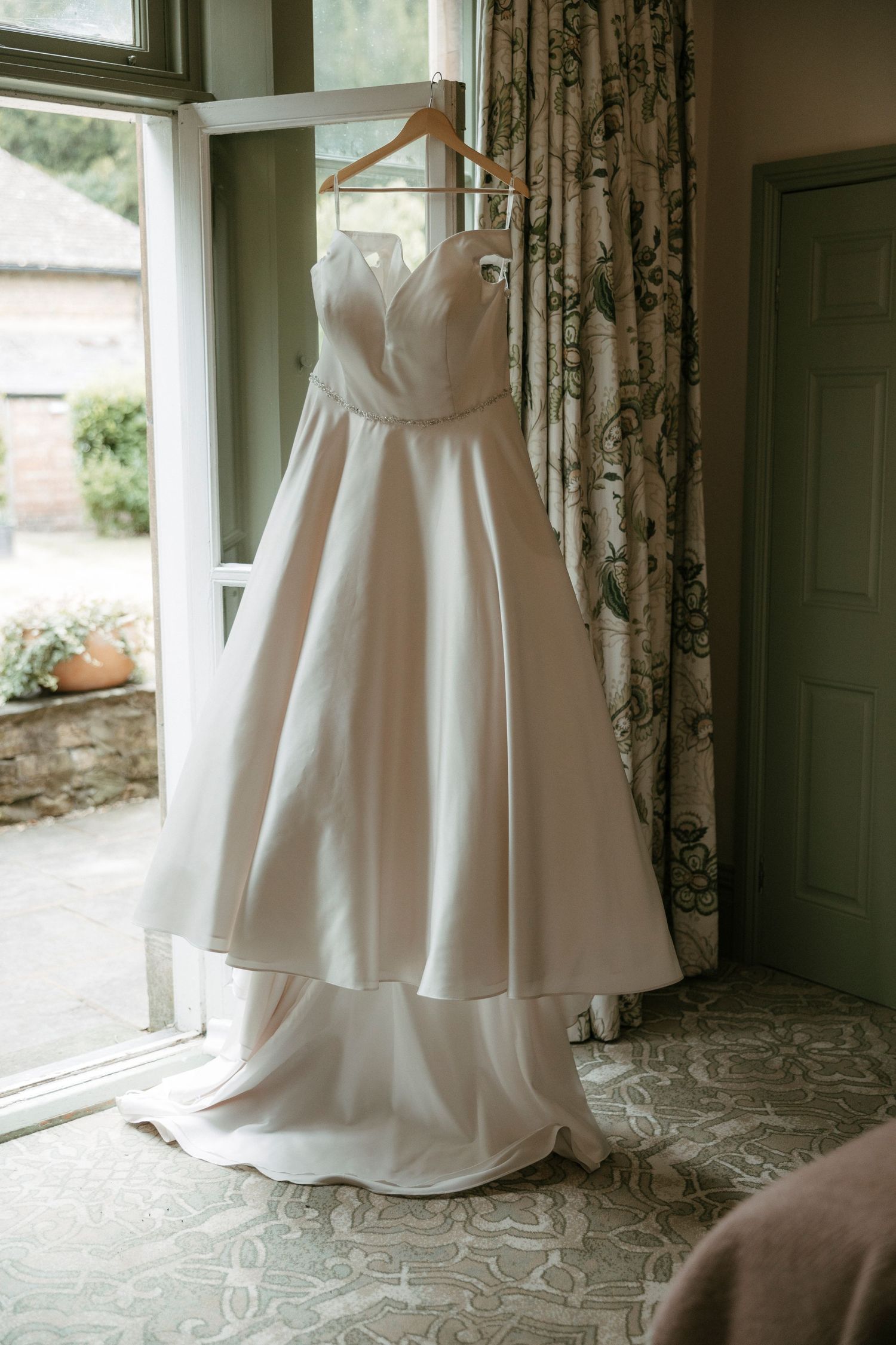 Elegant strapless ivory satin ball gown wedding dress hanging on wooden hanger by window with floral curtains.