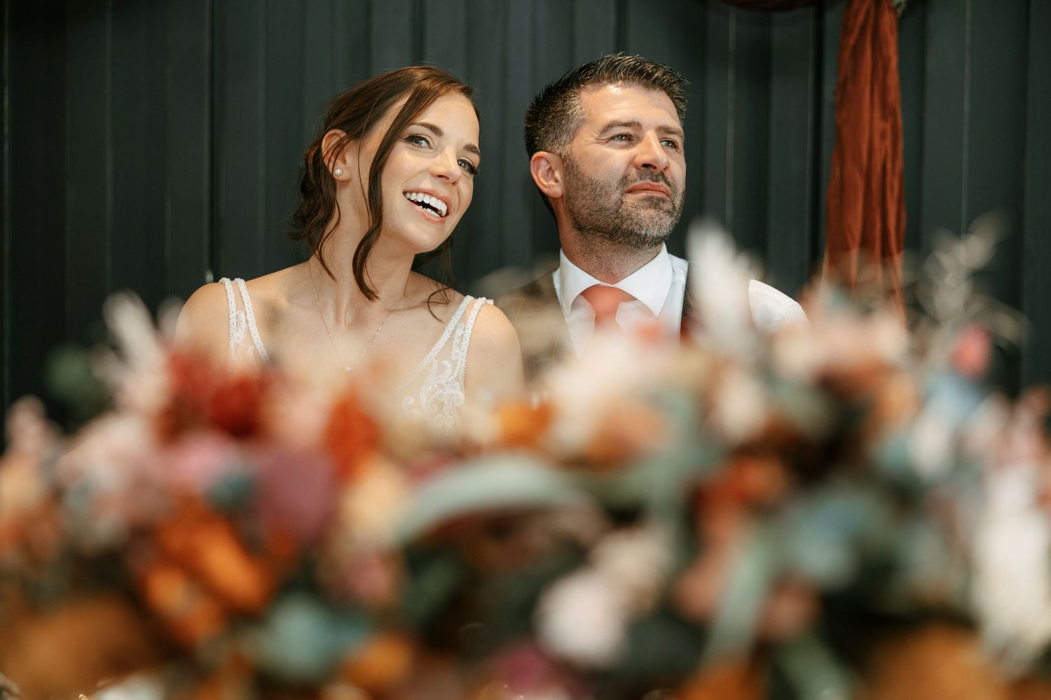 Happy bride and groom smiling at wedding ceremony with colorful floral bouquet in foreground.