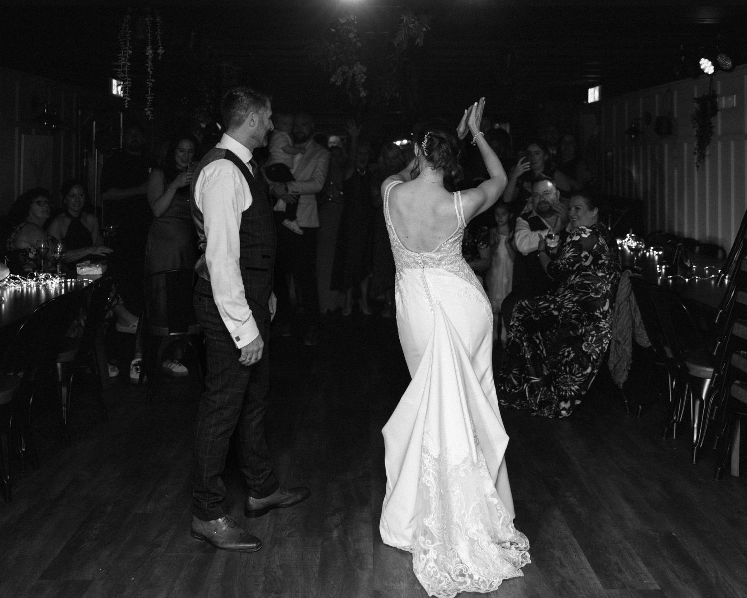 Black and white photo of bride and groom sharing first dance at wedding reception with guests in background.