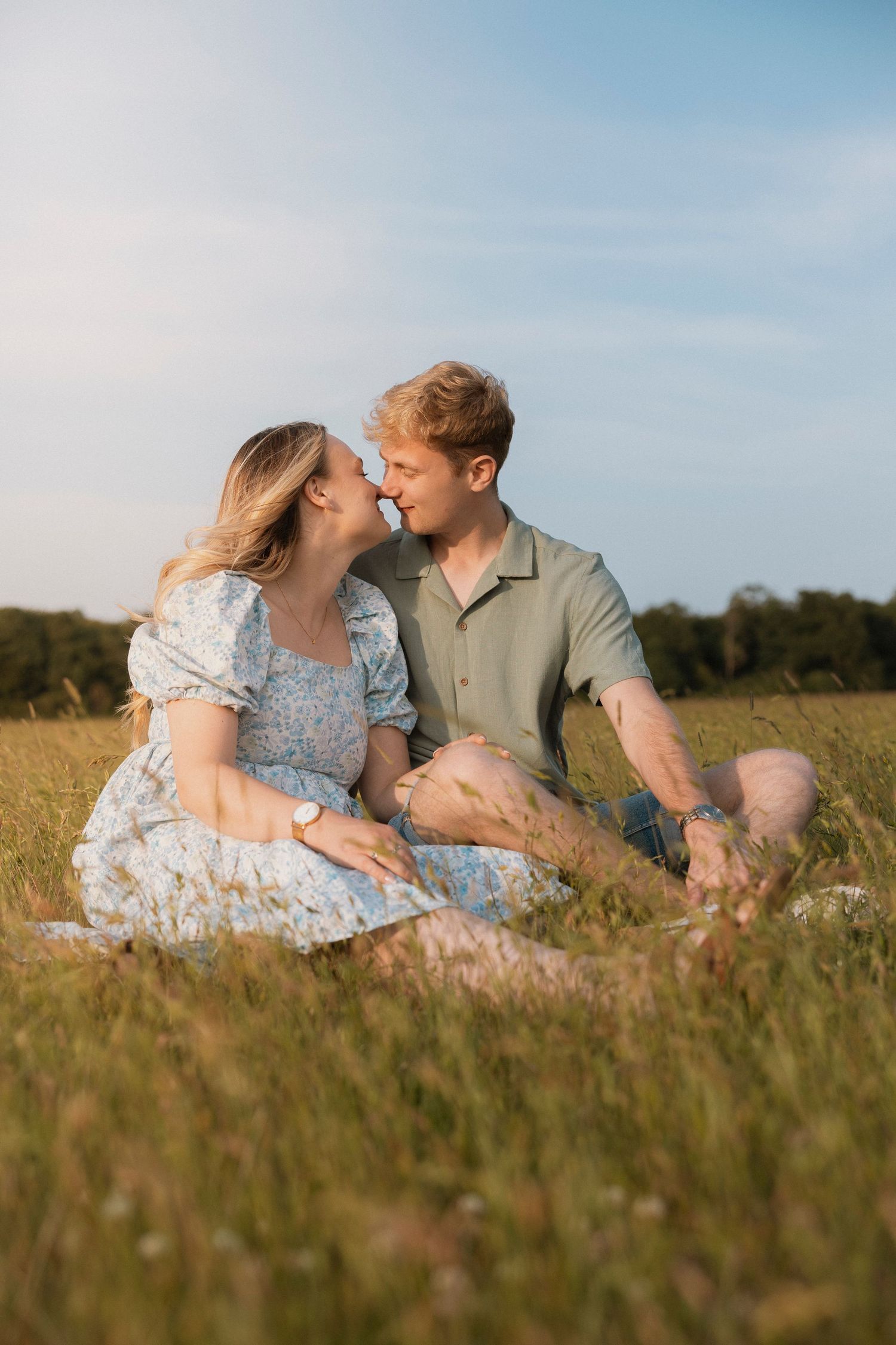 Couple sitting in a sunny green field, leaning in for a kiss, wearing casual summer outfits on a clear day.