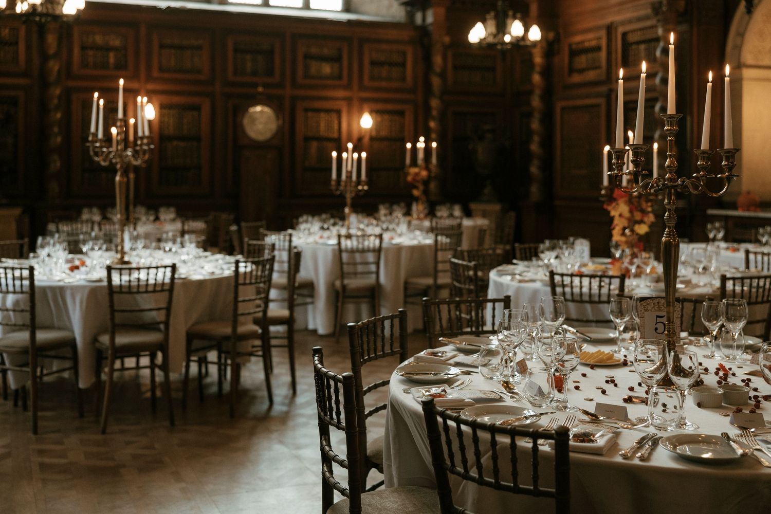 Elegant wedding reception hall with round tables, candelabras, and chiavari chairs in a wood-paneled venue.