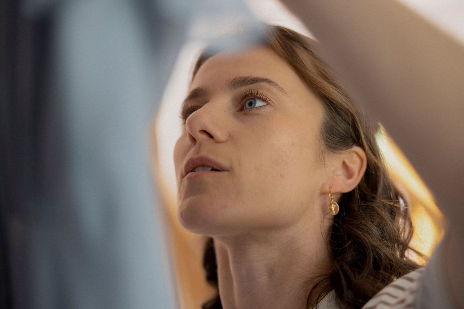 Close-up portrait of a young woman with brown hair and gold earrings gazing upward in soft golden light.
