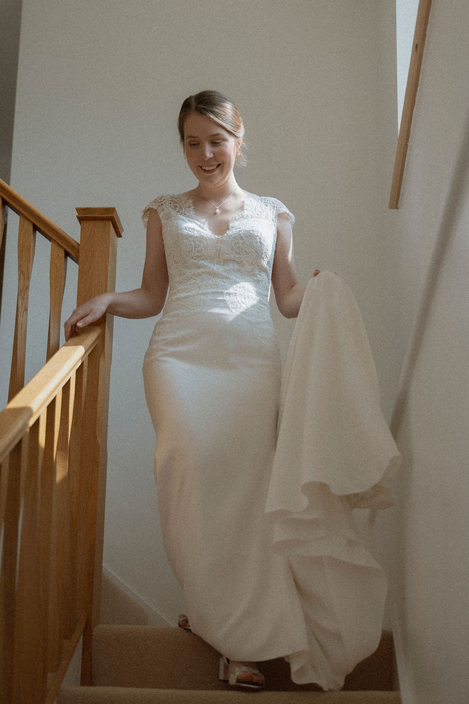 Smiling bride in a white lace wedding gown holding her train while descending wooden stairs on her wedding day.