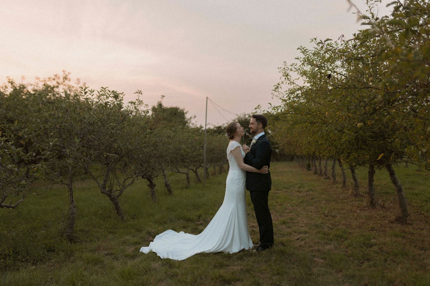 Bride and groom share a romantic moment in a lush orchard at dusk, bride in elegant white gown with long train.