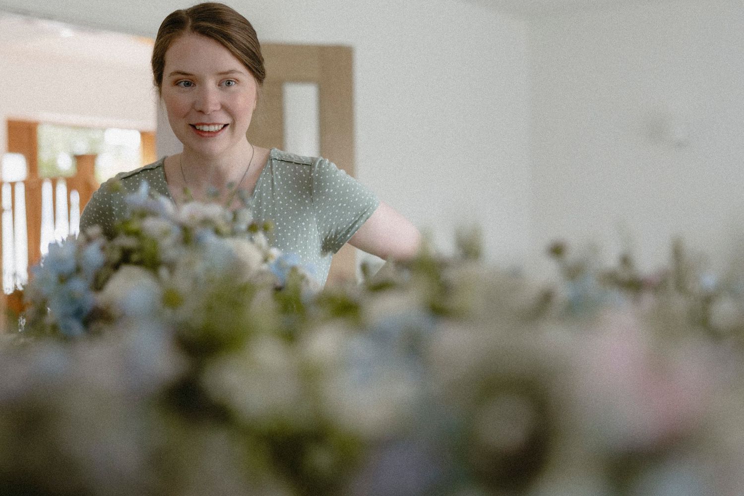Woman smiling while arranging pastel pink and blue flowers on a table in a bright room.